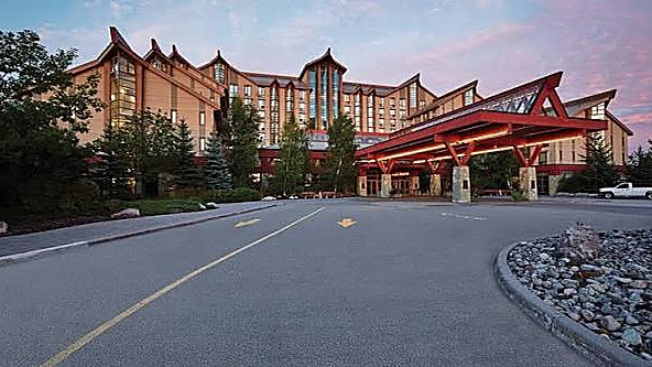 Hotel de madera y piedra con techos quebrados y una gran marquesina de acceso de vigas rojas, entre coníferas al amanecer.